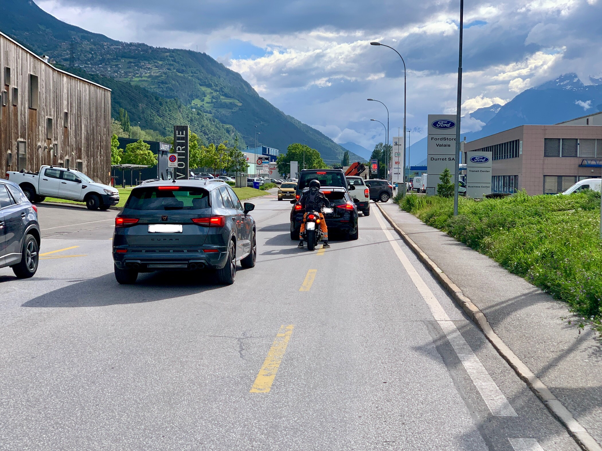 Une route à deux voies dans une zone montagneuse avec des voitures et des motos arrêtées dans la circulation. Des bâtiments et des concessionnaires automobiles bordent la route. Un ciel nuageux cache les sommets des montagnes en arrière-plan.