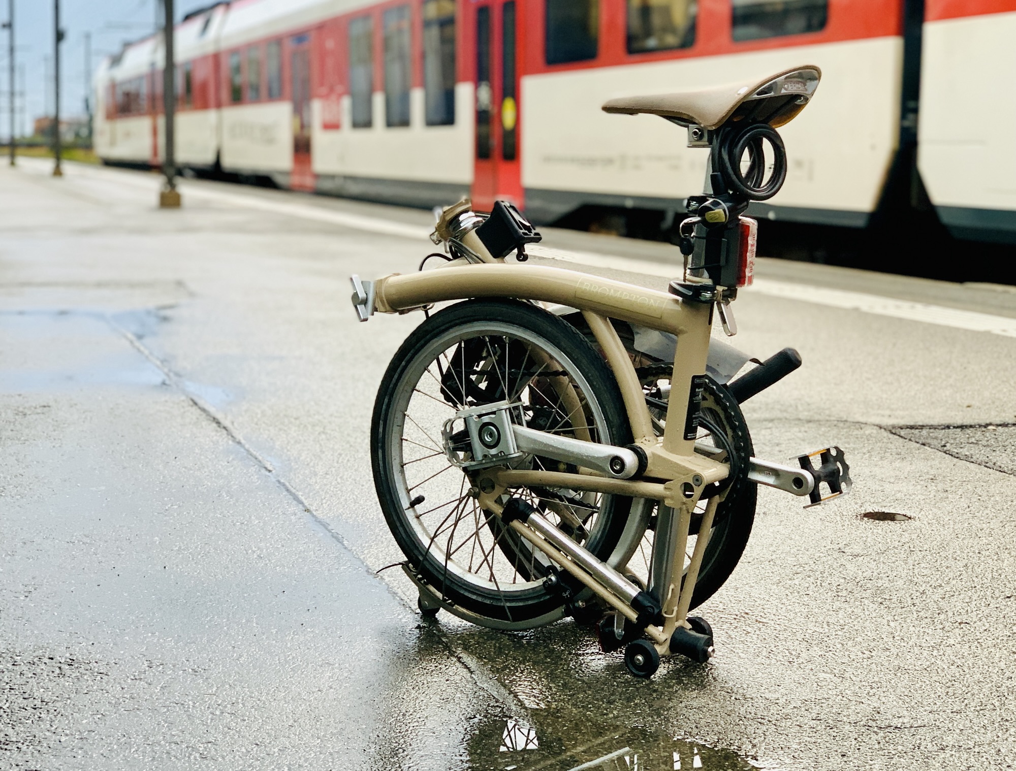 Vélo pliant beige compacté sur un quai de gare humide, avec un train rouge et blanc en arrière-plan. Idéal pour les déplacements urbains et les voyages en train.