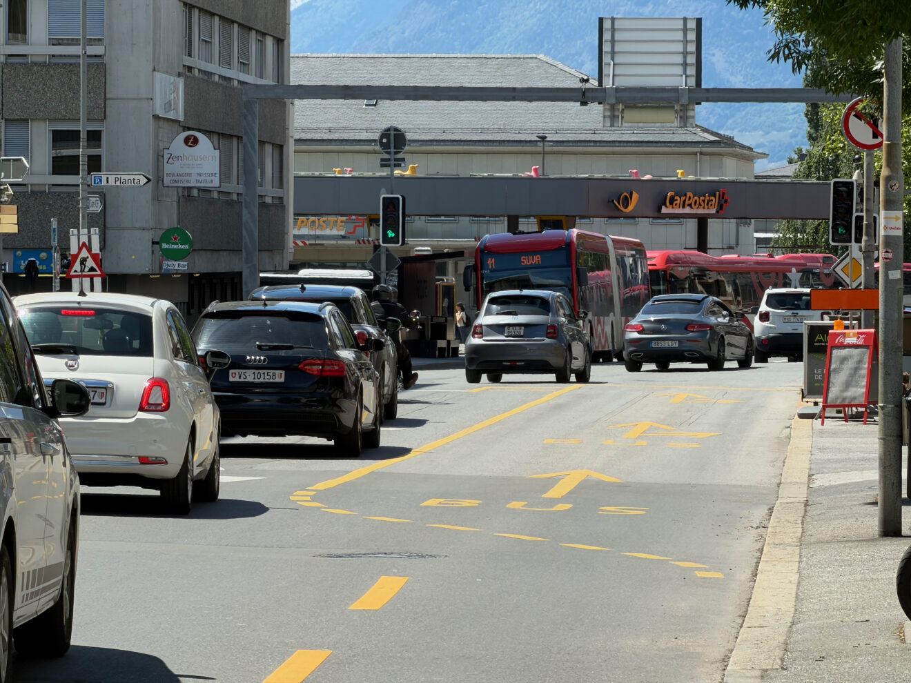 Une rue urbaine entourée d'immeubles, avec une voie de bus vide, avec de nombreuses autos sur la voie adjacente à un carrefour réglé par des feux de circulation, au vert. En face, des bus arrivent en sens inverse.