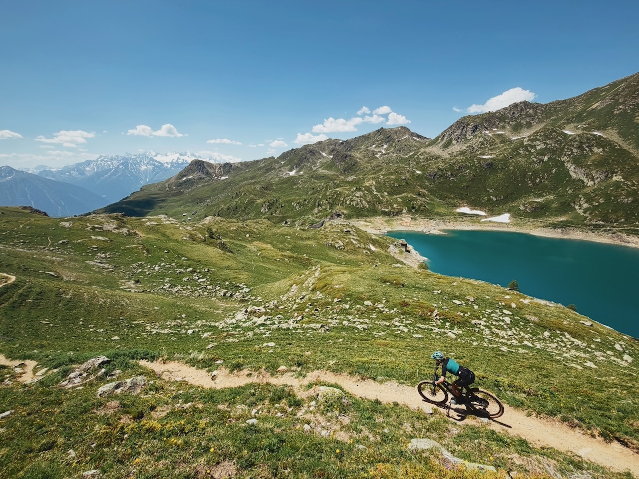 Cycliste en bleu sur une sentier de montagne, avec un lac bleu en contrebas et des montages enneigées à l'arrière-plan.