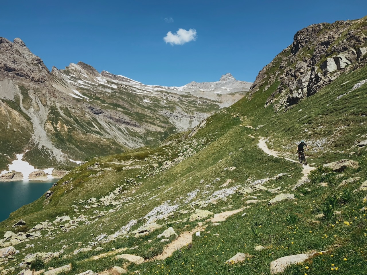 Paysage de montagne comprenant des sommets rocheux, un lac et une personne à vélo sur un sentier. Sur la droite, un chemin de terre serpente le long d'une pente verdoyante parsemée de rochers et de petites fleurs jaunes. Une personne circule à vélo sur ce sentier en direction du haut de la colline. À gauche, un lac bleu se trouve au pied de montagnes escarpées pré présentant des zones de neige ou de glacier. Le ciel est d'un bleu profond avec un petit nuage blanc isolé au centre.