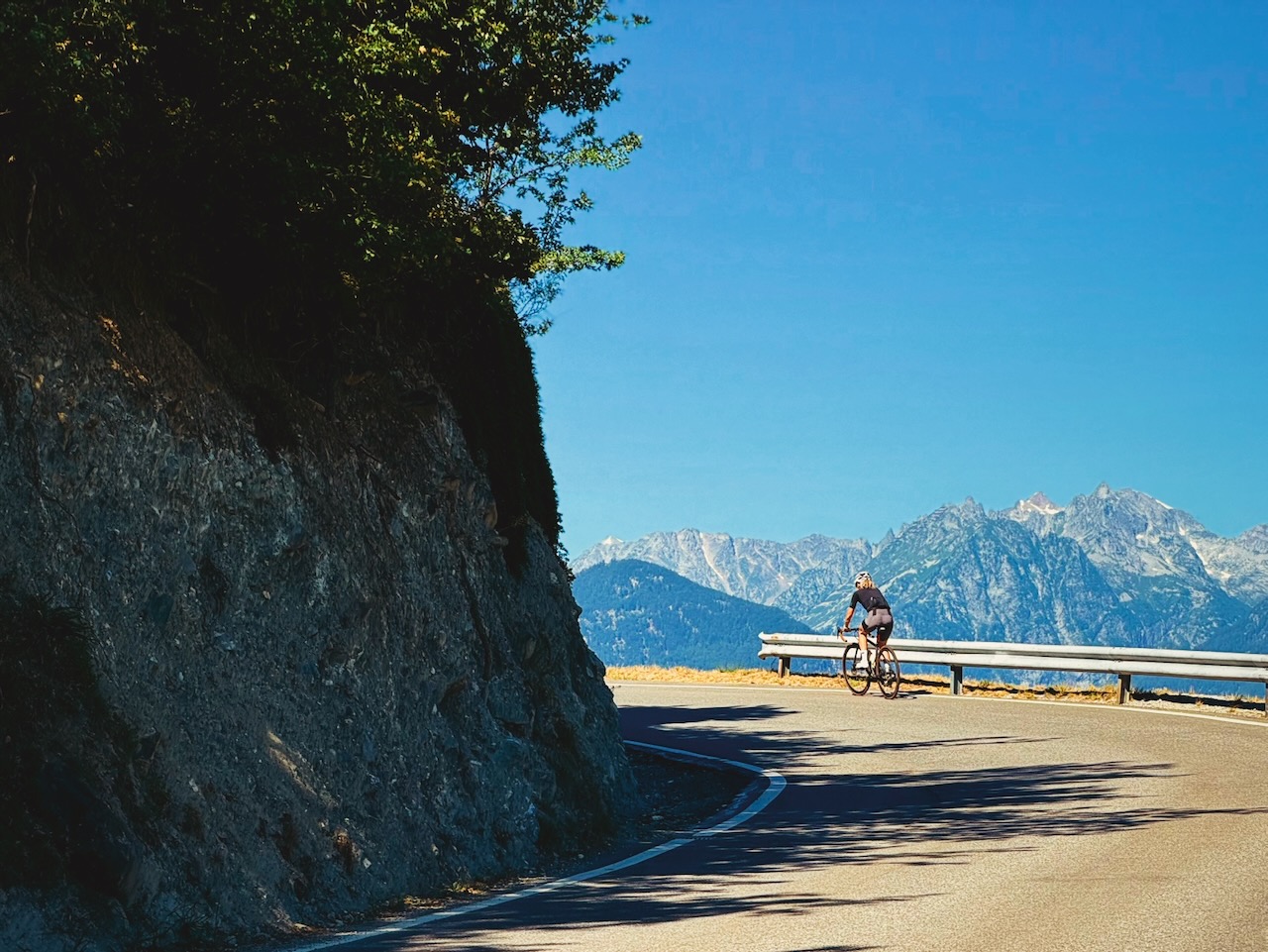Une cycliste sur une route de montagne, vue de l'arrière et à distance, en légère contre-plongée. Un rocher ombragé se dresse sur la gauche de la route, qui est au soleil avec un virage à gauche. La cycliste longe une glissière de sécurité, sur fond de montagnes sous un ciel bleu.