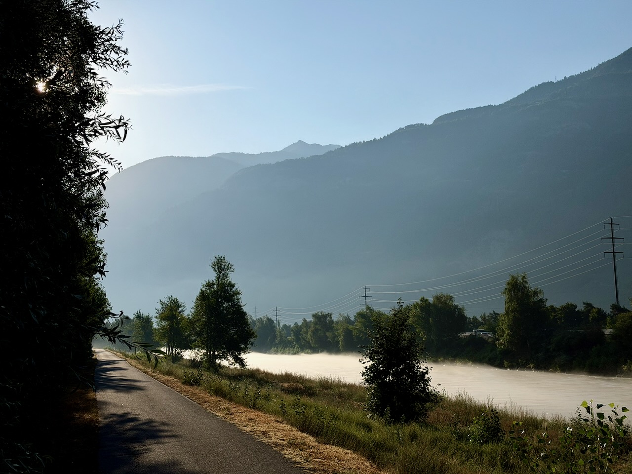 Une chaîne de montagnes derrière une vallée avec une rivière recouverte d'une brume blanche et basse. Au premier plan à gauche, des arbres en silhouette sombre cachent partiellement la vue, avec la lumière du soleil filtrant à travers les feuilles. Une route goudronnée part du coin inférieur gauche et mène vers la zone brumeuse. Le second plan montre des arbres et des buissons verts émergeant d'une épaisse couche de brouillard sur un champ. Des poteaux électriques avec des câbles sont visibles contre le pied des montagnes, qui s'estompent dans un bleu brumeux sous un ciel clair.