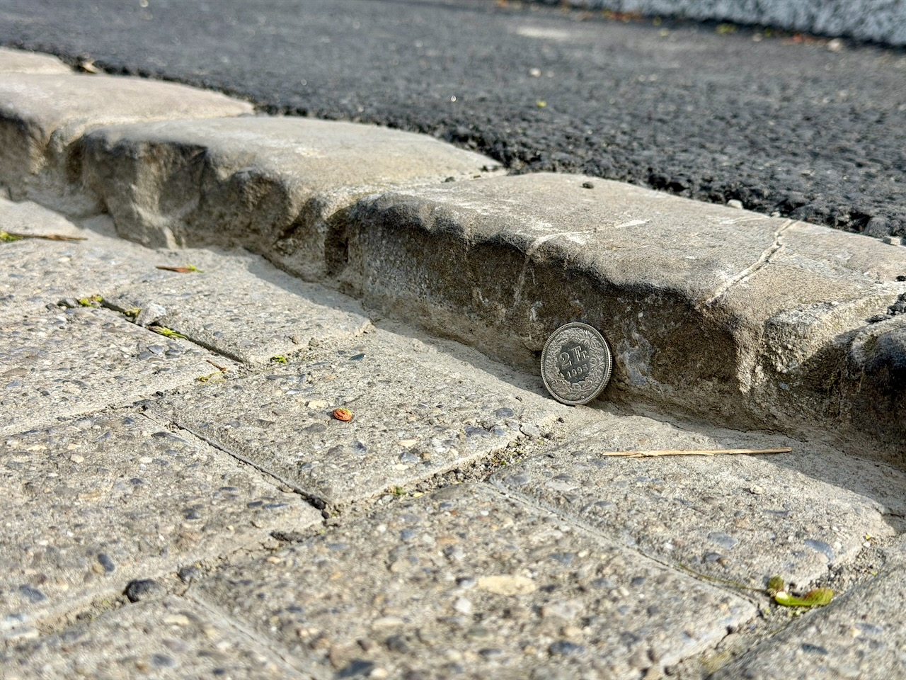 Gros-plan serré sur une bordure en pierre, deux fois plus haute qu'une pièce de deux francs suisses qui donne l'échelle.