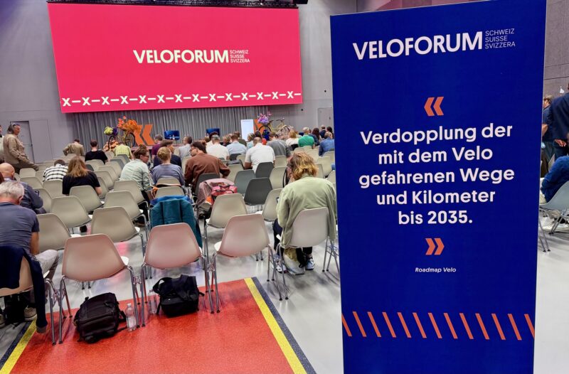 Vue d'une salle de conférence avec plusieurs rangées de chaises occupés, de dos. Sur scène, un affichage géant "Veloforum". Sur la droite, en premier plan, une affiche qui rappelle l'objectif de doubler la part modale du vélo d'ici 2035.