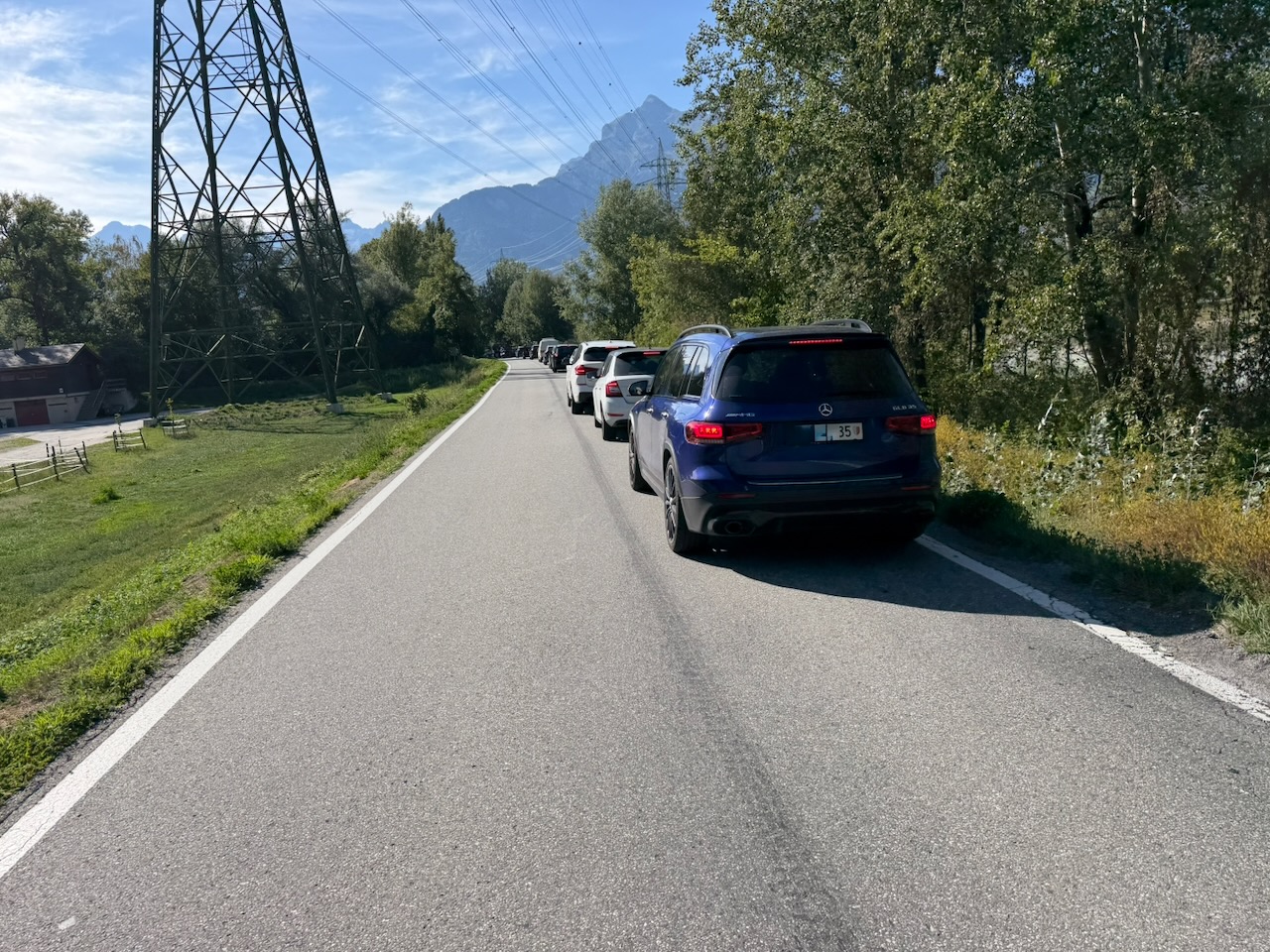 Une route avec une colonne de voitures sur la droite. Elle est entourée de verdure, avec des arbres à droite et un pylône sur la gauche. Au fond, on distingue des montagne sous un ciel légèrement voilé de nuages.