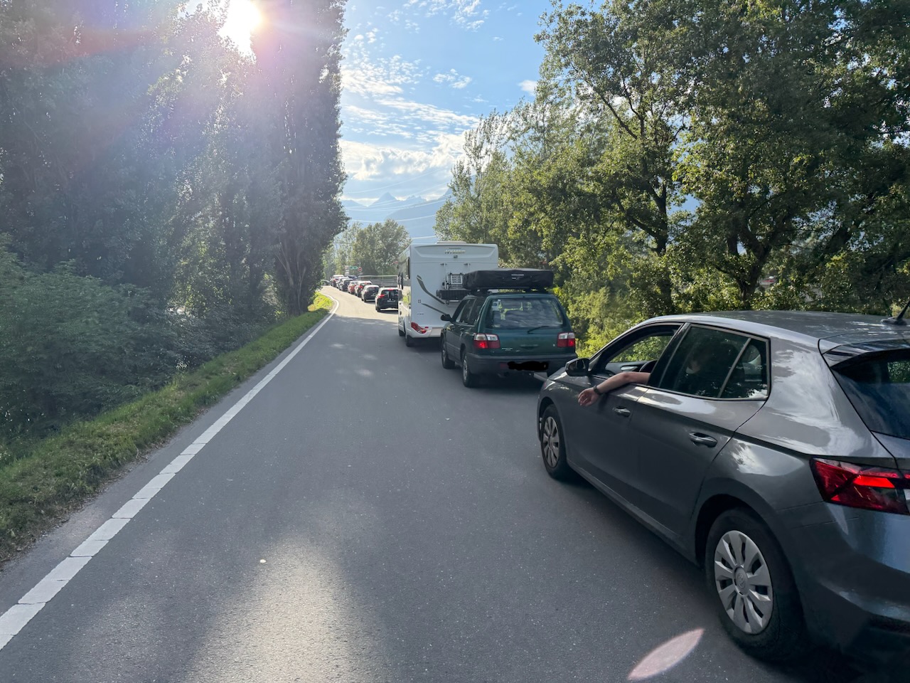 Une route avec une colonne de voitures sur la droite. Elle est entourée de verdure, avec des arbres à droite à gauche. On devine le soleil en coutre-jour, quelques montagnes dans le fond, sous un ciel bleu, légèrement voilé voilé.